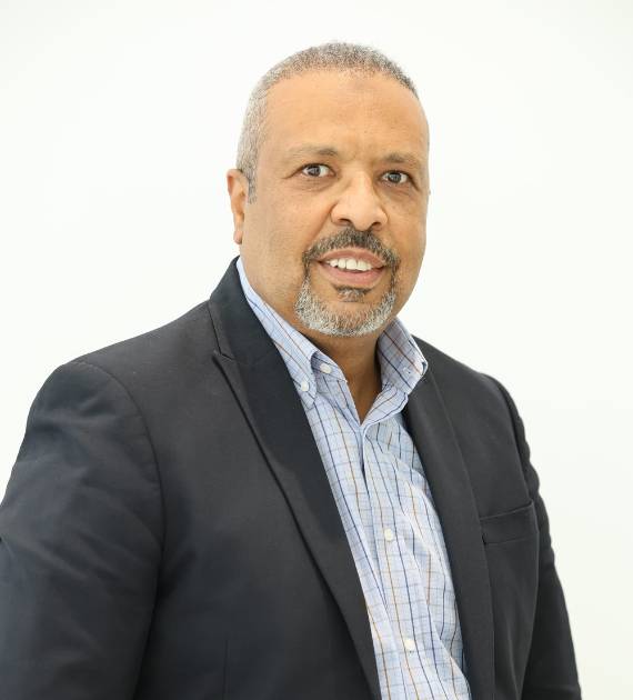 Hussein Fadloon, Project Director, Trojan Property Investment (TPI) - A professional man in a suit and blue checkered shirt smiling confidently against a plain background conveying expertise and approachability in a business environment.