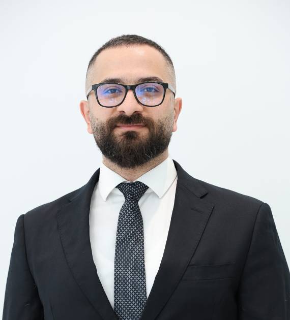 Yamen Almnini, Senior Architect, Trojan Property Investment (TPI) - A man in a formal dark suit and glasses with a neat beard stands against a plain light background exuding professionalism and style suitable for business environments.